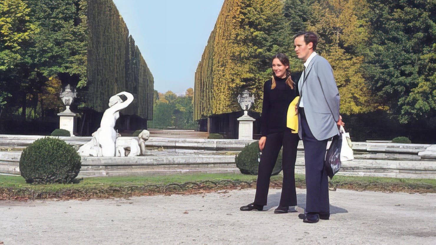 Two people - one in all black and one in a suit - stand in a well manicured garden. Behind them is a large head and some marble statues in a fountain.